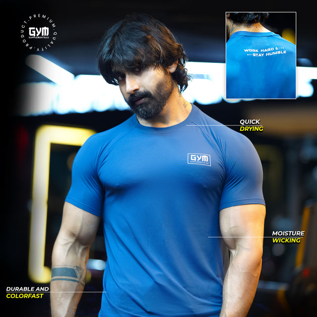 A muscular man wearing a blue Gym Supplements U.S. premium workout T-shirt featuring moisture-wicking and quick-drying fabric. The back shows the tagline "Work Hard & Stay Humble.