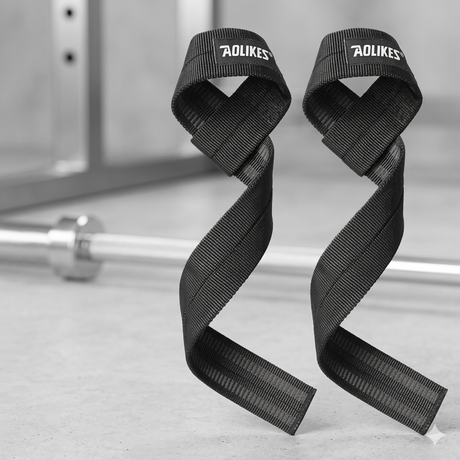 A pair of black AOLIKES cotton weightlifting lifting straps resting on a concrete gym floor next to a chrome barbell.