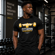 Muscular male fitness model standing in a gym wearing a black Gym Supplements U.S t-shirt featuring a yellow graphic of a bodybuilder squatting a heavy barbell above the word FITNESS.