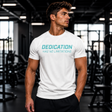 Muscular male fitness model standing in a gym wearing a white Gym Supplements U.S t-shirt featuring the teal text graphic "DEDICATION HAS NO LIMITATIONS".
